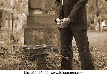 Stock Image - Man in a suit standing with folded hands at a gravesite. Fotosearch