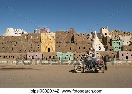 Stock Image - "Man on a motorcycle passing by the mud-brick model of the old town of Bawiti, Bahariya Oasis, Bawiti, Bahariya Oasis, Giseh, Egypt, Africa". Fotosearch