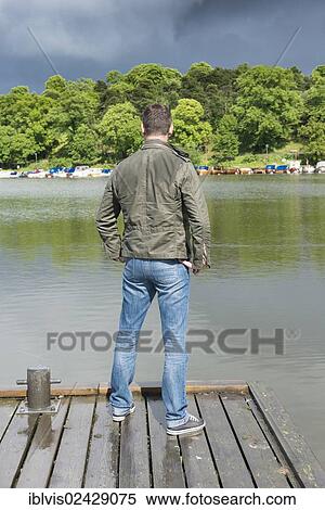 Stock Photography - Man standing on jetty by a lake and looking at rainclouds. Fotosearch