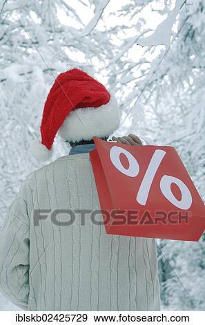 Stock Photo - "Man wearing a Santa hat in a snow-covered forest holding a shopping bag with a percent sign, rear view". Fotosearch