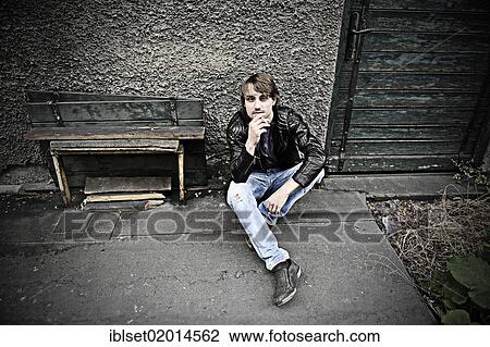 Stock Image - "Man wearing jeans and a leather jacket, holding a cigarette, sitting in front of an old wall, smoking". Fotosearch