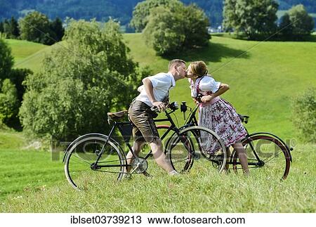 Stock Image - "Man wearing leather pants and a woman wearing a dirndl kissing on old bicycles within a natural landscape, Igls, Innsbruck, Tyrol, Austria, Europe". Fotosearch