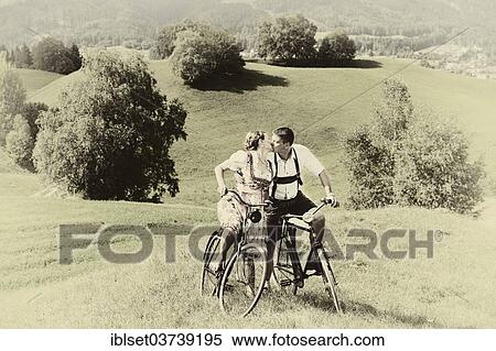 Stock Photography - "Man wearing leather pants and a woman wearing a dirndl kissing on old bicycles within a natural landscape, Igls, Innsbruck, Tyrol, Austria, Europe". Fotosearch