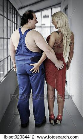 Stock Image - "Man wearing work clothes and a blonde woman wearing a red dress and high heels, grabbing each others buttocks". Fotosearch