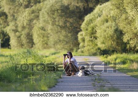 "Man with a dog crouching on a road and observing something with binoculars, Wustermark, Brandenburg, Germany, Europe" View Large Photo Image Stock Image - "Man with a dog crouching on a road and observing something with binoculars, Wustermark, Brandenburg, Germany, Europe". Fotosearch