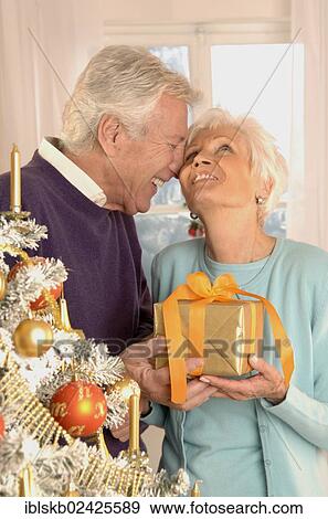 Mature couple holding a present next to a Christmas tree View Large Photo Image Stock Photo - Mature couple holding a present next to a Christmas tree. Fotosearch
