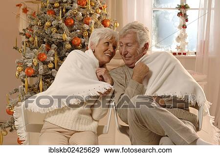 Stock Photo - Mature couple in love wrapped in a blanked sitting in front of a Christmas tree. Fotosearch