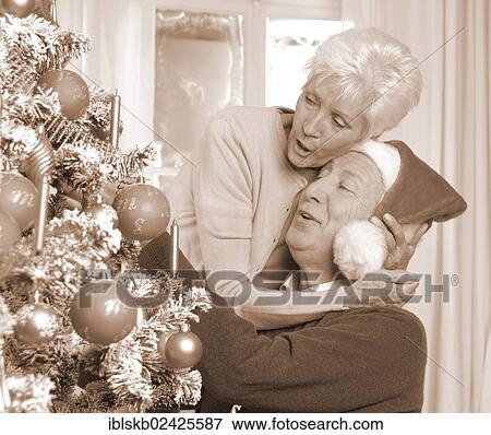 Stock Photo - "Mature couple, man with a Santa hat, by a Christmas tree". Fotosearch