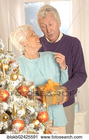 Stock Image - Mature couple with a present standing next to a Christmas tree. Fotosearch