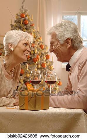 Stock Photo - Mature couple with red wine glasses and gift sitting in front of a Christmas tree. Fotosearch