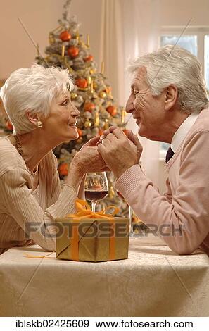 Stock Photo - Mature couple with red wine glasses and a present sitting in front of a Christmas tree. Fotosearch