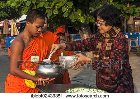 Stock Image - "Morning alms round, a young Buddhist monk from a monastery school holding a begging bowl, receiving rice or offerings from an older woman, Sukhothai Province, Northern Thailand, Thailand, Asia". Fotosearch