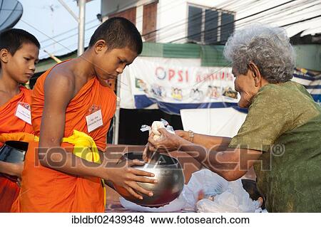 Stock Photo - "Morning alms round, a young Buddhist monk from a monastery school holding a begging bowl, receiving food or offerings from an older women, Sukhothai Province, Northern Thailand, Thailand, Asia". Fotosearch