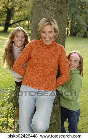Stock Photo - Mother and two daughters standing around a tree in a park. Fotosearch