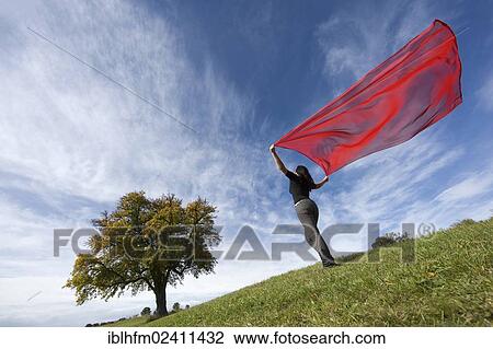 Stock Image - "Oblique side view of a young woman standing on a meadow, holding a red silk scarf fluttering in the wind, Aalen, Baden-Wurttemberg, Germany, Europe". Fotosearch