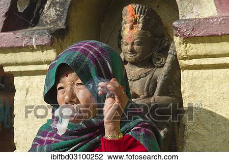 Stock Image - "Old woman, smoking, in front of a statue, Kathmandu, Kathmandu District, Bagmati Zone, Nepal, Asia". Fotosearch