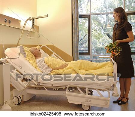 Stock Photo - Patient lying in a hospital bed is visited by a woman holding flowers. Fotosearch
