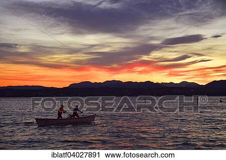 Stock Image - "People on a boat in Lake Garda, Lago di Garda, with sunset, Desenzano del Garda at the back, Sirmione, Lombardy, Italy, Europe". Fotosearch