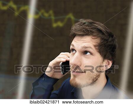 Stock Image - Portrait of a young businessman with a beard making a telephone call, relaxed, smiling, successful, focused, share prices graph at back. Fotosearch