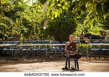 Stock Image - "Praying or meditating Buddhist monk sitting on a plastic chair, temple and monastery of Wat Phra Archa Thong or Golden Horse Temple, Mae Chan, Chiang Rai Province, Northern Thailand, Thailand, Asia". Fotosearch