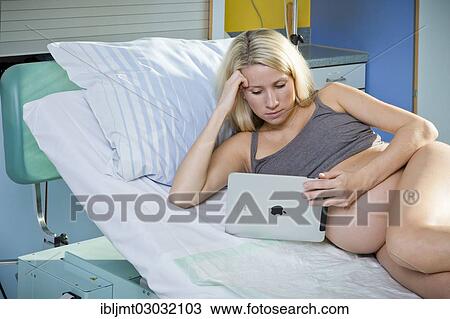 Stock Image - "Pregnant woman lying in the delivery room on the delivery bed and looking at a tablet PC, Germany, Europe". Fotosearch