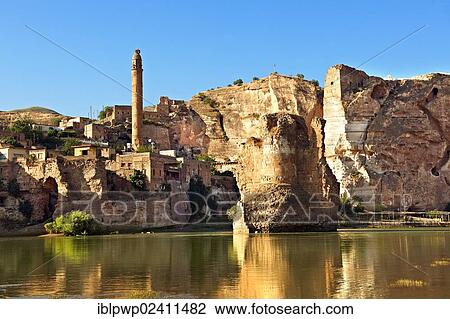 Stock Image - "Remains of medieval Artukid, old Tigris bridge, built in 1116 by Artukid Fahrettin Karaaslan, with the old town Hasankeyf and its ruins on the cliffs abover the river Tigris and the minaret of the El Rizk Mosque, built in 1409, Turkey, Asia". Fotosearch