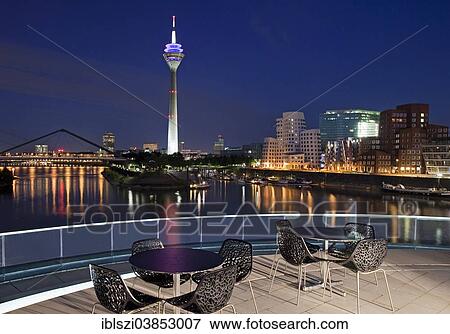 Stock Photo - "Restaurant terrace with Rheinturm tower and the Gehry buildings at the blue hour, Neuer Zollhof, Media Harbour, Dusseldorf, North Rhine-Westphalia, Germany, Europe". Fotosearch