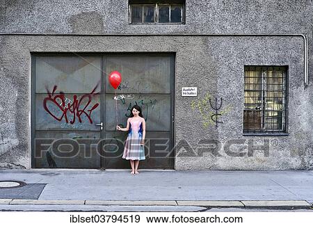 Stock Photo - Sad girl holding a red balloon in front of an old garage door. Fotosearch