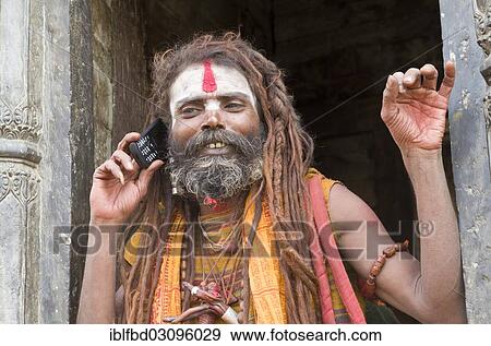 Stock Photo - "Sadhu, Holy Man, using his mobile phone opposite the burning ghats, Kathmandu, Kathmandu District, Bagmati Zone, Nepal, Asia". Fotosearch