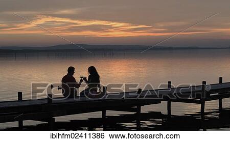 Stock Photograph - "Silhouette of a couple in the twilight sitting on a wooden pier on the lake toasting with red wine glasses, Steinhude am Meer, Lower Saxony, Germany, Europe". Fotosearch