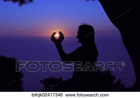 Stock Photograph - Silhouette of a woman against the sunset sky, her hands enclosing the setting sun, Lefkas, Greece, Europe. Fotosearch