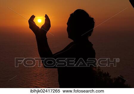 Stock Photography - Silhouette of a woman against the sunset sky, her hands enclosing the setting sun, Lefkas, Greece, Europe. Fotosearch