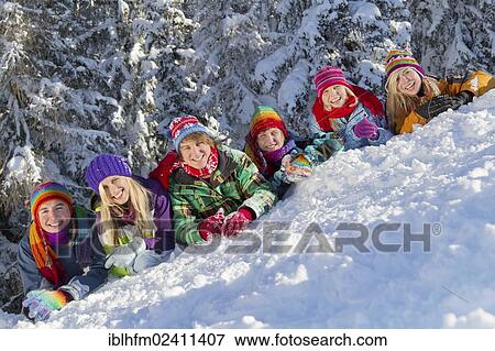 Stock Photo - "Six smiling young people lying on the snow slope in front of a snow-covered forest, Flachau, Salzburg, Austria, Europe". Fotosearch