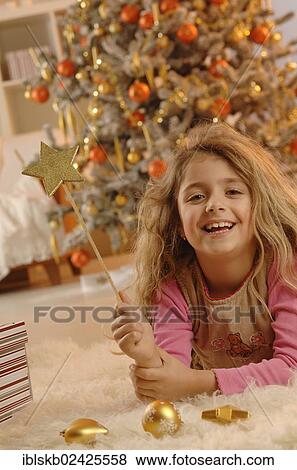 Stock Photo - Smiling girl lying on the ground with a golden star and gifts in front of a Christmas tree. Fotosearch
