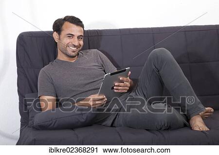 Smiling man lying on a gray sofa with an iPad View Large Photo Image Stock Image - Smiling man lying on a gray sofa with an iPad. Fotosearch