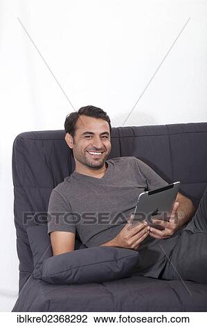 Stock Image - Smiling man lying on a gray sofa with an iPad. Fotosearch