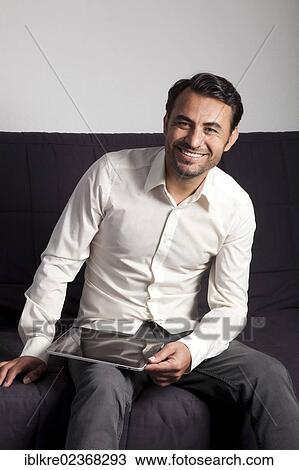 Smiling man sitting on a sofa with an iPad View Large Photo Image Stock Image - Smiling man sitting on a sofa with an iPad. Fotosearch