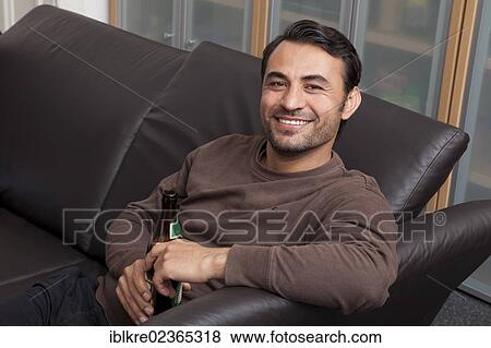 Smiling man sitting with a bottle of beer on a leather sofa View Large Photo Image Stock Photo - Smiling man sitting with a bottle of beer on a leather sofa. Fotosearch