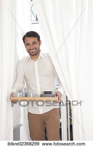 Smiling man standing at a balcony door with curtains and carrying a tray of dishes View Large Photo Image Stock Photo - Smiling man standing at a balcony door with curtains and carrying a tray of dishes. Fotosearch