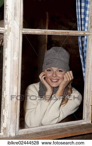 Stock Photo - Smiling young woman wearing a hat and a scarf sitting at the window of a hiking hut. Fotosearch
