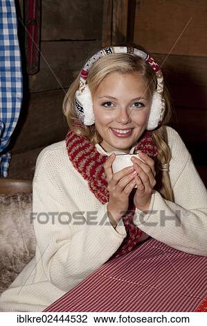 Smiling young woman wearing ear warmers and a scarf sitting at the table of a hiking hut and drinking a hot drink View Large Photo Image Stock Image - Smiling young woman wearing ear warmers and a scarf sitting at the table of a hiking hut and drinking a hot drink. Fotosearch