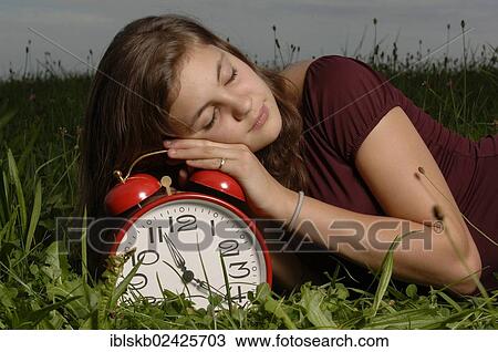Stock Image - Teenage girl lying on the grass and sleeping on an oversized alarm clock. Fotosearch