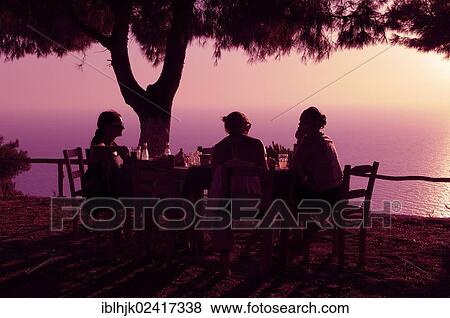 Three women sitting at a table in a seaside garden at sunset, Lefkas, Greece, Europe View Large Photo Image Stock Photo - Three women sitting at a table in a seaside garden at sunset, Lefkas, Greece, Europe. Fotosearch