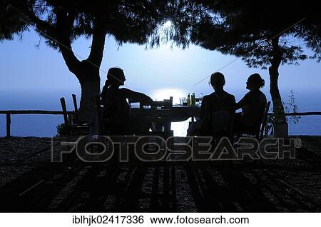 Three women sitting at a table in a seaside garden at sunset, Lefkas, Greece, Europe View Large Photo Image Stock Photograph - Three women sitting at a table in a seaside garden at sunset, Lefkas, Greece, Europe. Fotosearch