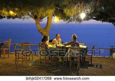 Three women sitting at a table under a tree, in a Greek seaside garden in the evening, Lefkas, Greece, Europe View Large Photo Image Stock Photo - Three women sitting at a table under a tree, in a Greek seaside garden in the evening, Lefkas, Greece, Europe. Fotosearch