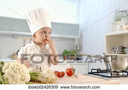 Stock Photo - "Toddler, 2-4, wearing a chef's hat in a kitchen and eating a tomato". Fotosearch