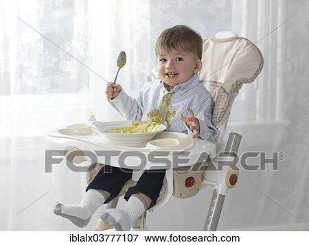 Toddler Boy Sitting In A High Chair Eating Soup With A Spoon