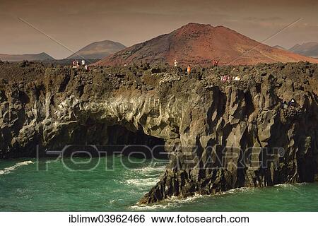 Stock Photograph - "Tourists on the rocky coast of Los Hervideros, volcanic landscape, the Fire Mountains of Timanfaya National Park at the back, Lanzarote, Canary Islands, Spain, Europe". Fotosearch
