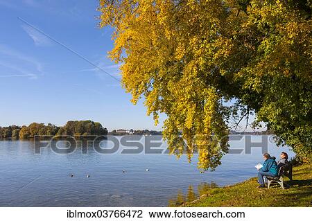 Stock Image - "Tourists sitting on a bench at Lake Chiemsee, Island Frauenchiemsee or Fraueninsel, Chiemsee, Upper Bavaria, Bavaria, Germany, Europe". Fotosearch