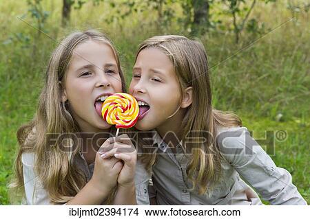 Picture - "Twin girls, 9, licking a colourful lollipop". Fotosearch